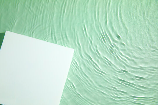 Empty white square podium on transparent clear green calm water texture with splashes and waves in sunlight. Abstract nature background for product presentation. Flat lay cosmetic mockup, copy space.