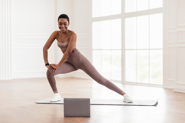 Young black woman doing side lunges on mat using laptop