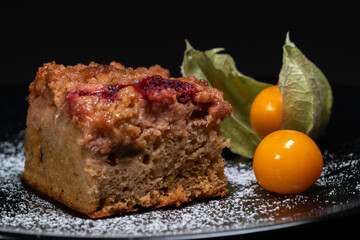Closeup shot of a freshly baked cake with raspberries, jostaberries and rhubarb on a black plate isolated on black background with two ripe physalis.