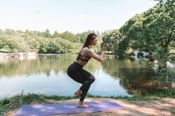 Young attractive woman in sportswear practicing yoga,standing in Eagle Pose, Garudasana,in the park on the lake shore.Sports healthy lifestyle.Outdoor training.Slow living concept.Copy space.