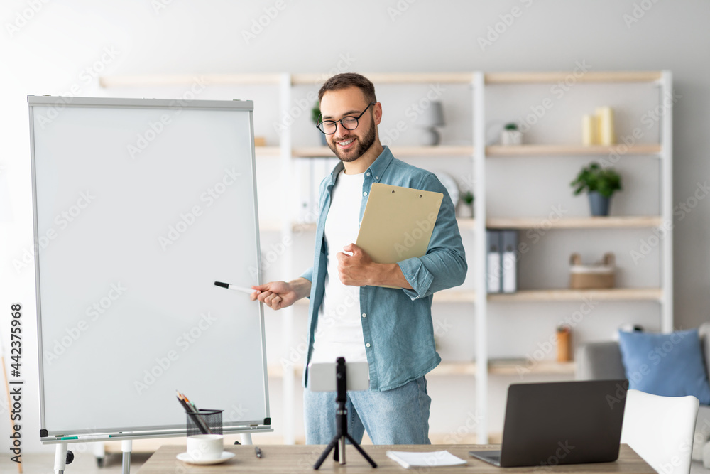 Poster young male teacher pointing at empty blackboard during web lesson, using smartphone and laptop at ho