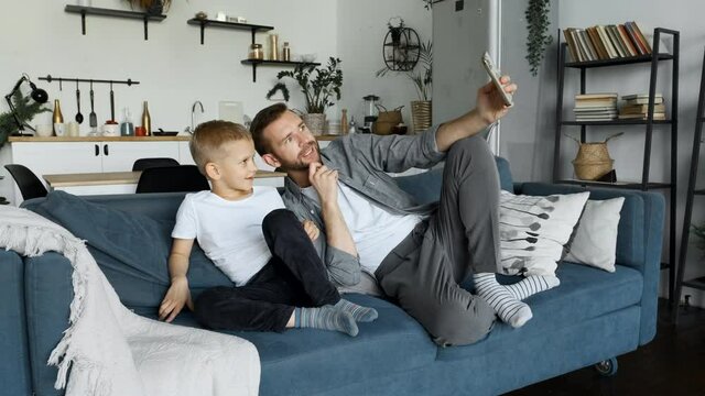 Father and child son waving hands looking at camera, make video call with digital smartphone with family while sitting in the living room. Their are laughing relaxing with video chatting with his fami