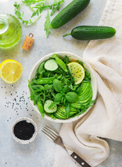 Green vegan (vegetarian) bowl-salad of fresh vegetables-arugula, cucumber, avocado, green peas, coriander, basil with lemon juice, olive oil and black sesame on a gray background. Healthy food, diet