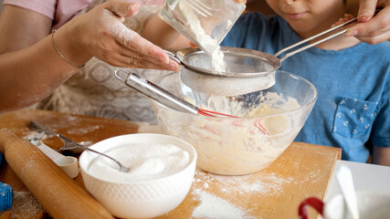 Closeup of little boy with mother sifting flour with sieve. Children cooking with parents, little chef, family having time together, domestic kitchen
