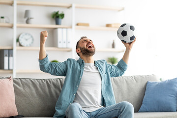 Emotional guy watching soccer on TV, lifting ball above his head and shouting, celebrating goal or...