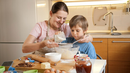 Young smiling mother teaching her son sifting flour with sieve for making biscuit dough. Children cooking with parents, little chef, family having time together, domestic kitchen.