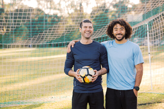 Two men smiling as they stand together in a soccer  goal, with one of the men holding a soccer ball