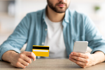 Shopping at home. Closeup of young guy holding credit card and smartphone, making online purchase at table indoors