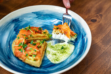 waffle with salmon and white sauce on blue plate is eaten with a knife and fork.