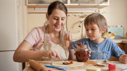 Smiling mother with little son filling cake or pie with sweet jam.Children cooking with parents, little chef, family having time together, domestic kitchen