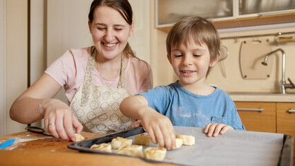 Smiling boy with mother making biscuits and putting them on baking sheet. Children cooking with parents, little chef, family having time together, domestic kitchen.