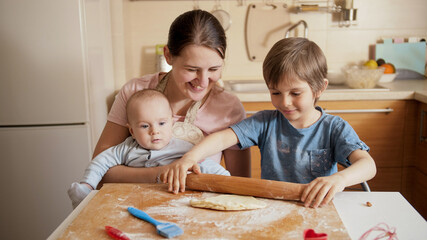 Young mother with little baby son looking at her older son rolling dough and cooking. Children cooking with parents, little chef, family having time together, domestic kitchen.