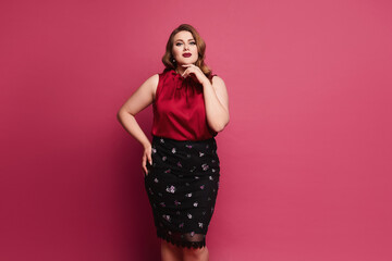Young plus size caucasian model woman in red satin blouse and skirt posing over pink background. Gorgeous plump woman in modish outfit over pink background
