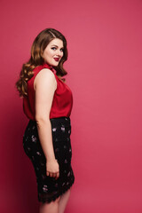 Young plus-size model girl in red satin blouse and skirt posing over pink background. Happy chubby woman in modish outfit over pink background