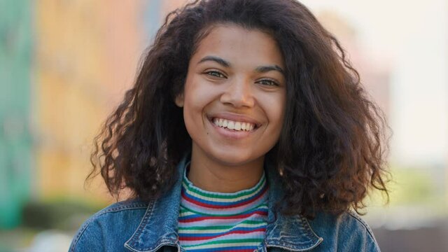 Portrait of Smiling Afro-American Girl Student with Curly Hair standing outdoors. Attractive Young Lady wears Casual Clothes looking into camera having a Good Mood. Ethnic Appearance. Beauty Female.
