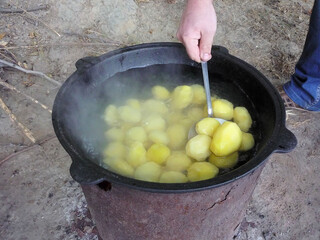 Whole peeled potatoes are cooked in water in a black cauldron on the street, a person's hand stirs...