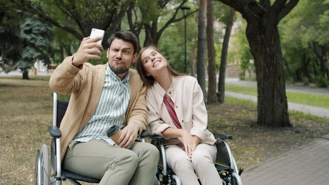 Joyful disabled couple man and woman are taking selfie with smartphone camera posing with funny faces sitting in wheelchairs in park having fun together.