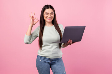 Photo of charming positive young woman smile show okay sign hold laptop isolated on pink color...