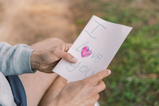A letter with an I love you text and a red heart written on a small paper held by a man in two hands