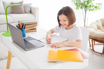 Portrait of attractive cheery focused diligent girl attending lesson watching video tutorial writing essay at light white flat home indoors