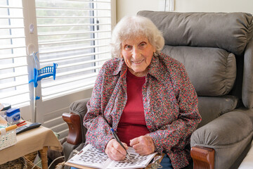 An elderly lady sitting in a chair doing a crossword and smiling
