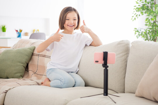 Full body photo of young little girl happy positive smile speak video call cellphone show thumbs-up cool fine choice sit couch indoors