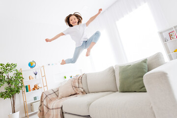 Full size photo of young excited small girl happy positive smile have fun jump up fly air sofa indoors