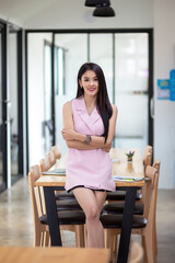 Asian businesswoman in a pink dress stands with her arms crossed looking at the camera at the office.