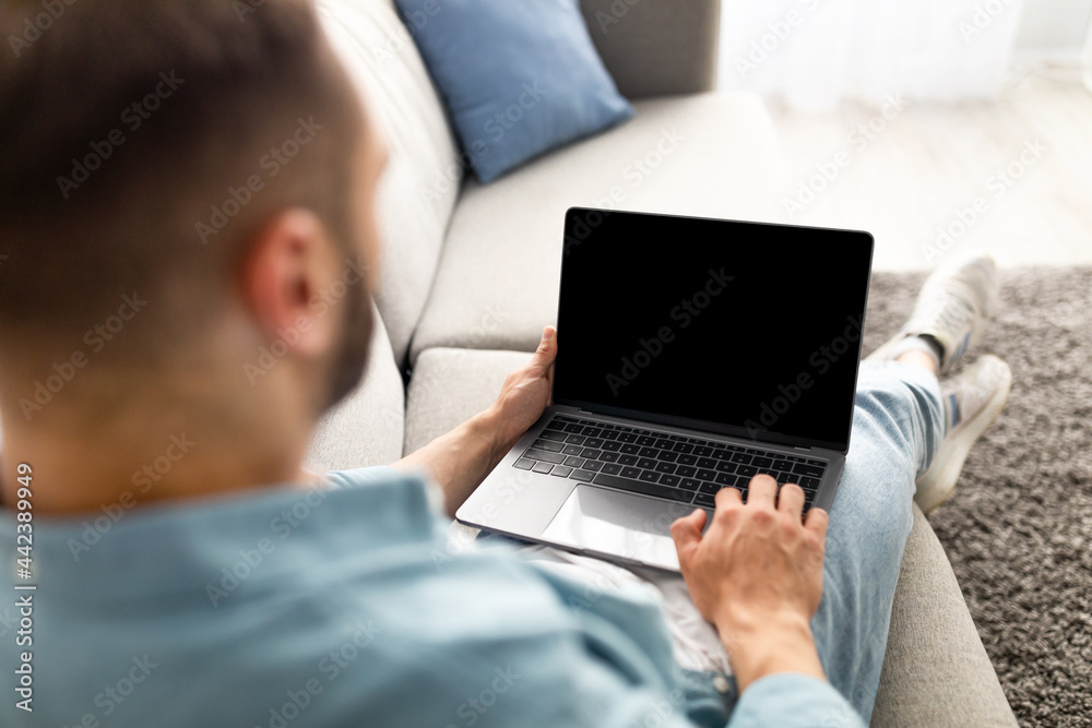 Poster unrecognizable young man using laptop on comfy sofa at home, mockup for website design on screen
