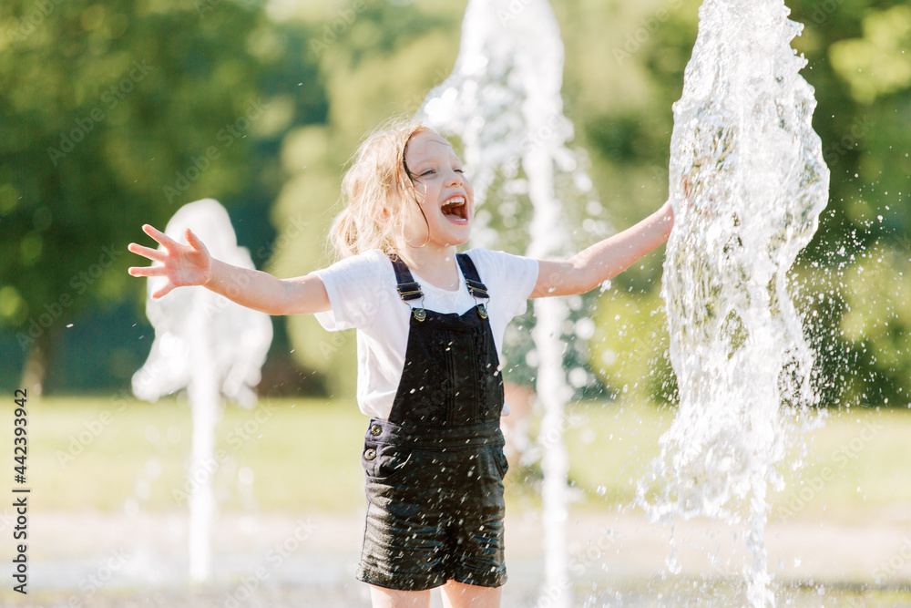 Wall mural Cute cheerful girl playing in fountain. Kid in denim overall having fun in summer park