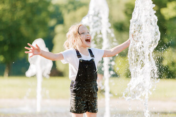 Cute cheerful girl playing in fountain. Kid in denim overall having fun in summer park