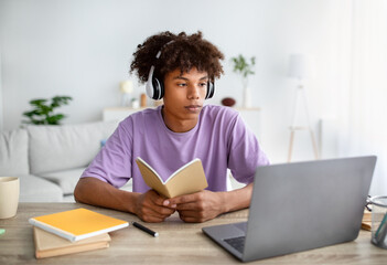Intelligent black teen student in headphones studying online in front of laptop, holding notebook at home