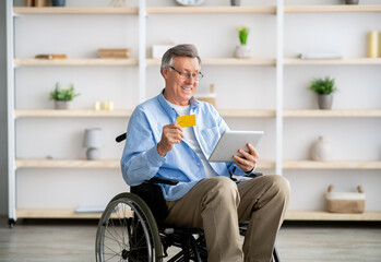Positive disabled older man in wheelchair shopping online with credit card and tablet pc at...