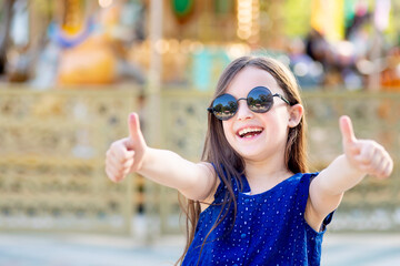 a child girl in an amusement park in the summer shows the class the upper finger up and smiles with...