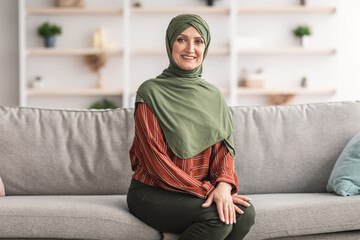 Happy Mature Muslim Woman Wearing Hijab Sitting On Sofa Indoors