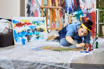 Talented female painter in apron choosing paint color, holding paintbrush while working on...