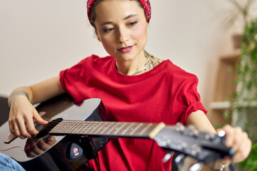 Young woman in casual clothes looking focused while playing guitar at home