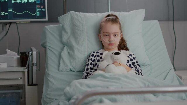 Portrait of lonely little child looking into camera holding teedy bear in hands resting in bed suffering infection. Kid waiting for recovery treatment during medical examination in hospital ward