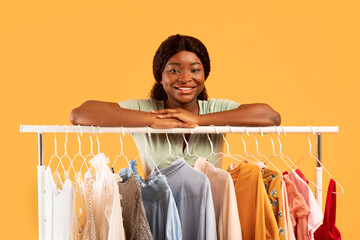 Cheerful black female stylist leaning on clothing rack, smiling at camera, offering choice of trendy summer outfits