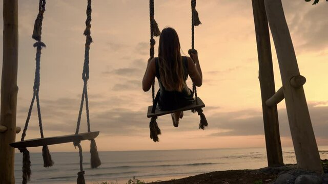 A dark silhouette of a girl on swing at seaside. A beautiful vacation leisure on a beach at sunset near ocean shore. Tropical romance and enjoyable time swinging alone.Anonymous female flying on swing