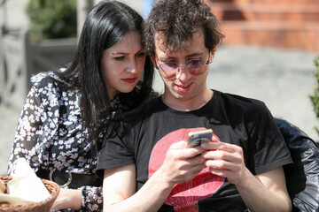 The relationship of a married couple. The man uses his smartphone and is happy. The woman looks over his shoulder and gets upset.