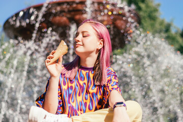 Happy pink-haired teenage hipster girl in a colorful bright T-shirt is eating ice cream on a summer...