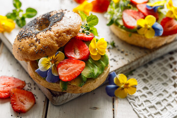 Close up view of a vegan bagel sandwich with edible pansy flowers, strawberries and avocado. A...