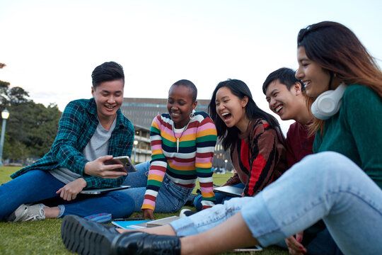 Group of young university students hanging out sitting on grass studying and using devices