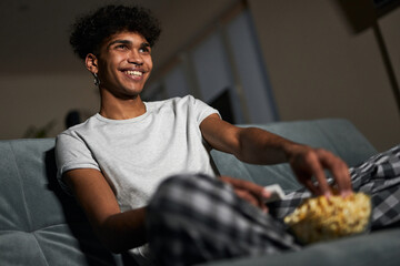 Cheerful young guy wearing pajamas taking some popcorn from a bowl while watching comedy, sitting...