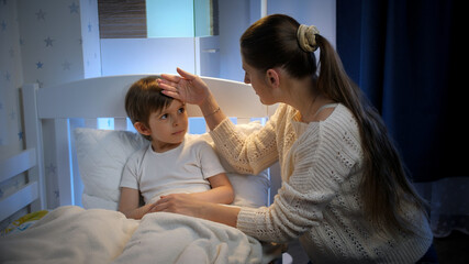 Young mother sitting next to her sick little son lying in bed at night. Concept of children illness, disease and parent care