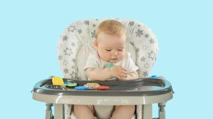 Baby in the feeding chair front view. A six-month-old baby is sitting on a high chair playing with toys and pulling them into his mouth