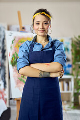 Smiling young woman, female artist in apron looking at camera, standing with her arms crossed at home studio workshop