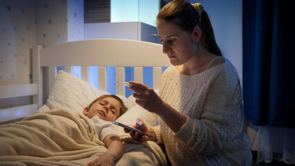 Young mother calling doctor or 911 emergency to her sick little son lying in bed. Concept of...