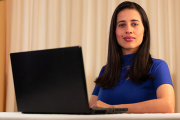 Fototapeta premium Woman sitting in front of her advertising type laptop, looking at the camera. young man, student, promoting the virtual environment. Latin woman, single and white skin. For Advertising.